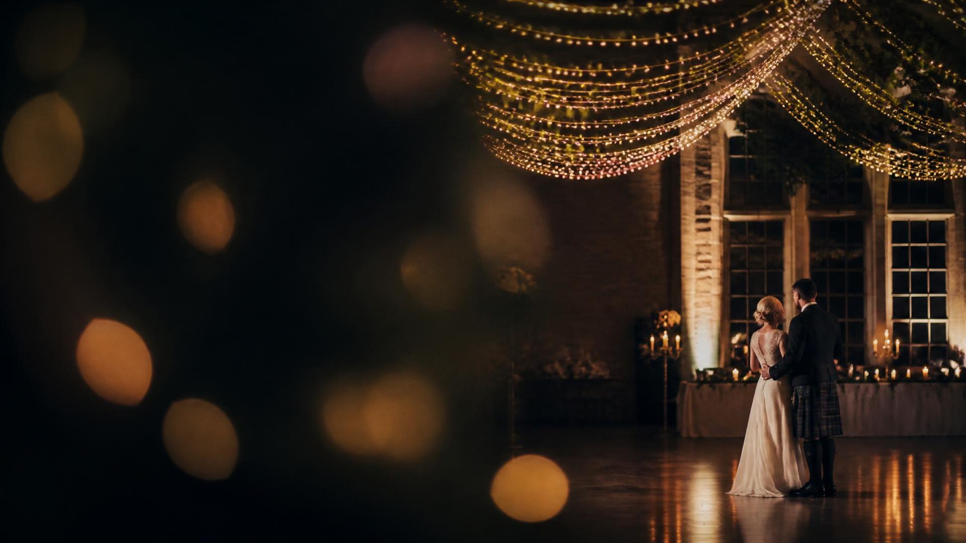 Bride and groom in a kilt dancing together in a candlelit country-house ballroom beneath a fairy-light canopy
