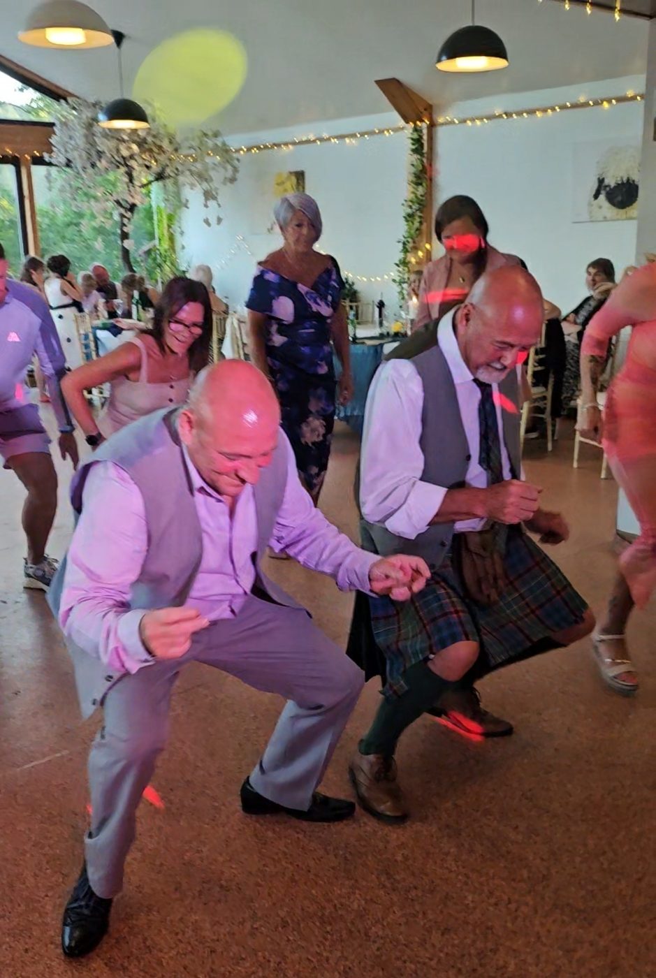 Guests in kilts dancing at a daytime Scottish wedding reception with fairy lights overhead