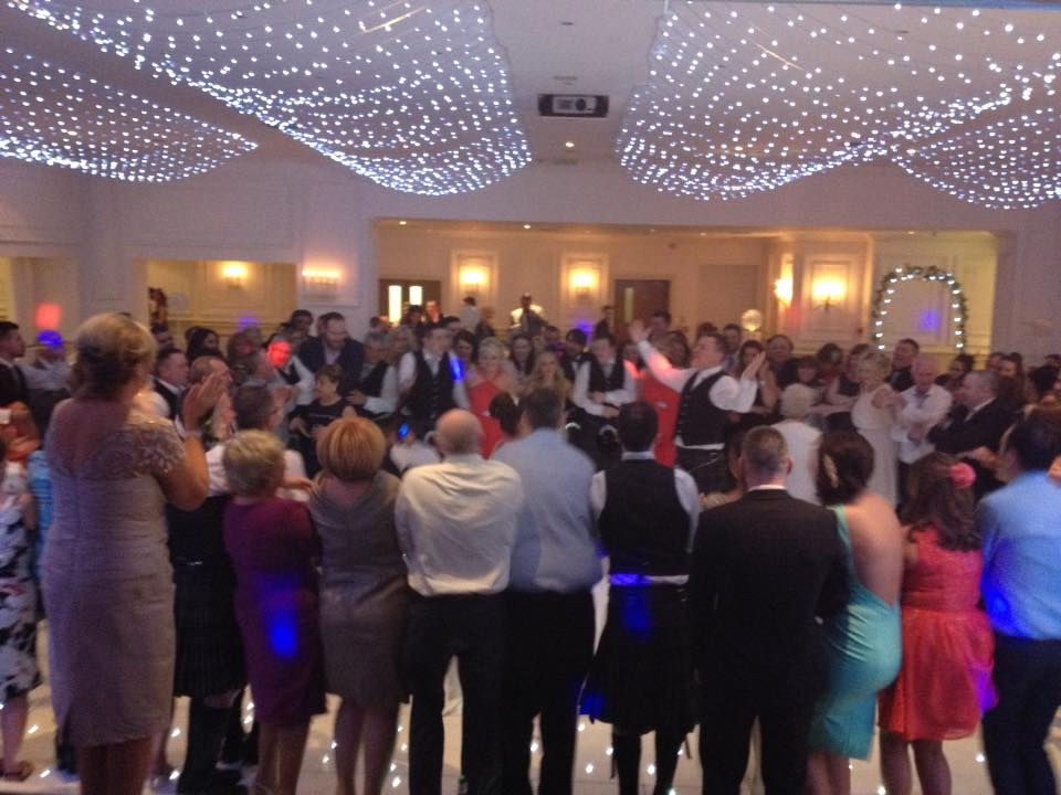 Wedding guests gathered in a circle clapping and singing in a function room lit by fairy-light drapes