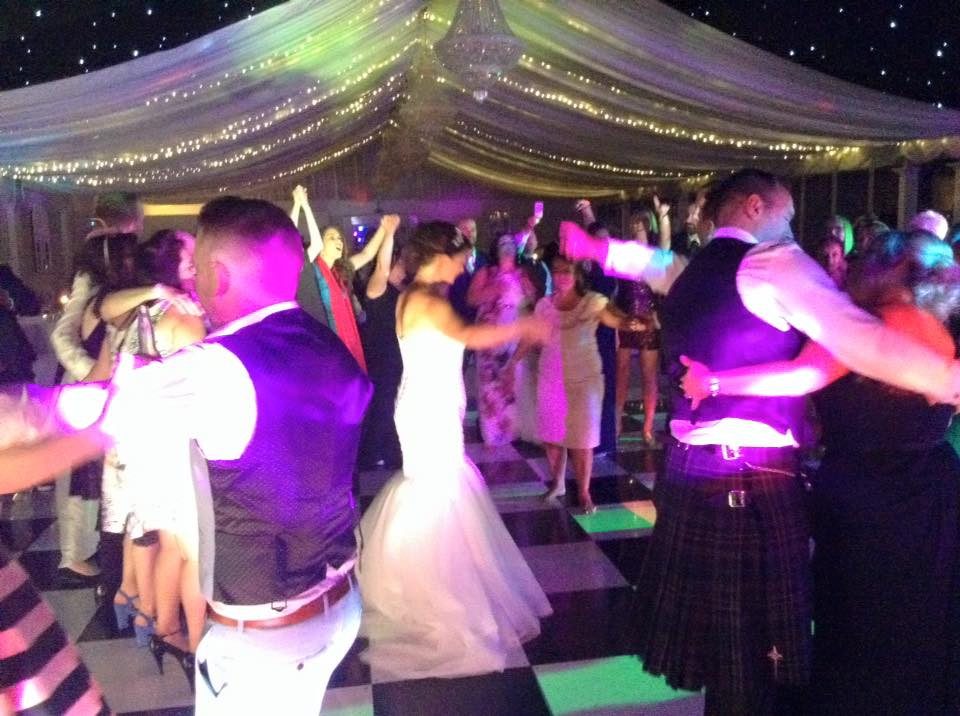 Bride and guests in kilts dancing on a chequered dancefloor beneath a draped fairy-light canopy at a Scottish wedding
